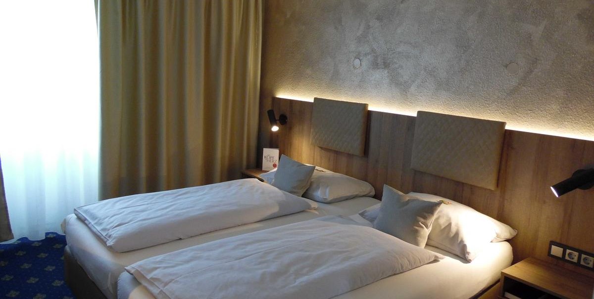 Modernes Doppelzimmer mit zwei Einzelbetten, Nachttischlampen und einem Fenster mit Vorhängen. Die Wand ist mit Holzpaneelen verkleidet., © Hotel Arcis Modernes Doppelzimmer mit zwei Einzelbetten, Nachttischlampen und einem Fenster mit Vorhängen. Die Wand ist mit Holzpaneelen verkleidet., © Hotel Arcis