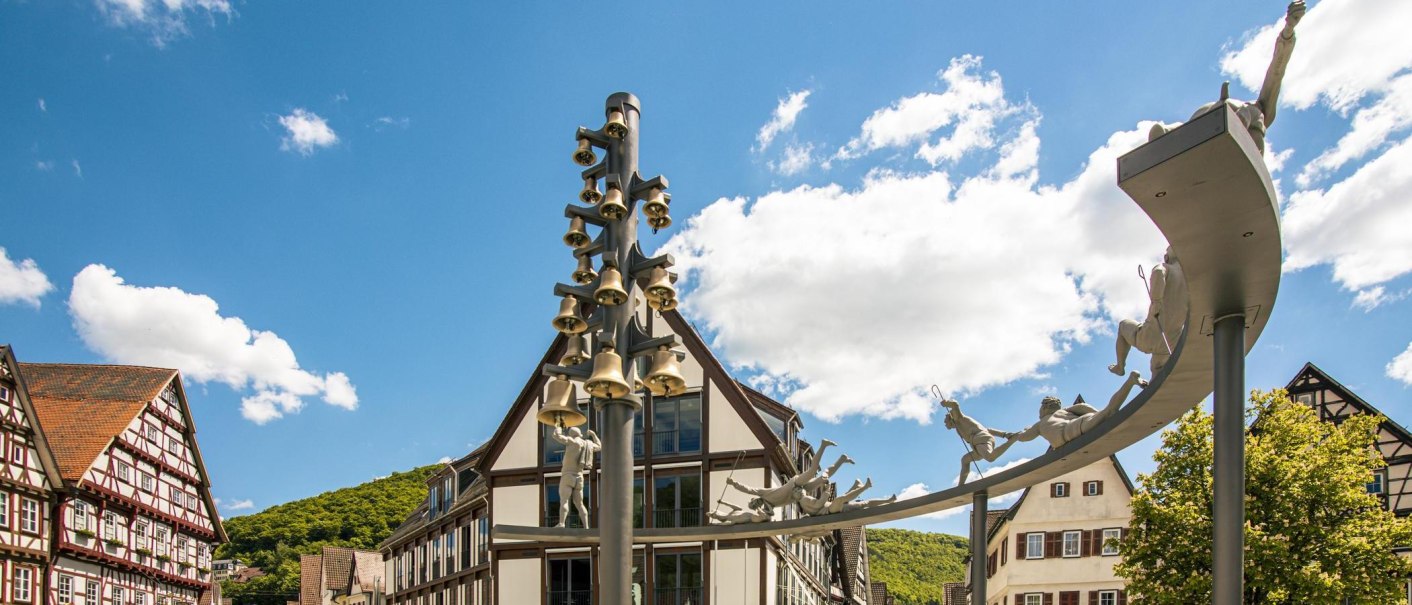 Skulptur mit Glocken und Figuren auf dem Marktplatz von Bad Urach, umgeben von Fachwerkhäusern und blauem Himmel., © Stuttgart-Marketing GmbH, Sarah Schmid Skulptur mit Glocken und Figuren auf dem Marktplatz von Bad Urach, umgeben von Fachwerkhäusern und blauem Himmel., © Stuttgart-Marketing GmbH, Sarah Schmid