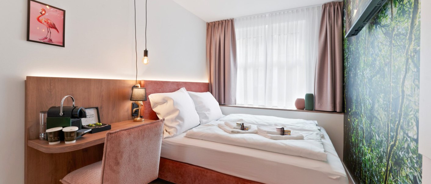 Modernes Zimmer mit Doppelbett, rosa Akzenten, Wandbild eines Waldes und Flamingo-Bild. Schreibtisch mit Kaffeemaschine und stilvoller Beleuchtung., © JUNGLE N°5 GmbH