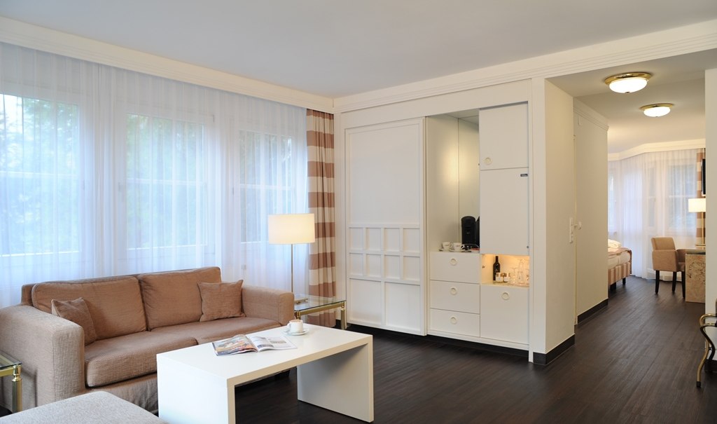 Heller Wohnbereich einer Junior Suite mit beigem Sofa, weißem Couchtisch und Blick auf den Schlafbereich. Große Fenster mit Vorhängen., © relexa Waldhotel Schatten Heller Wohnbereich einer Junior Suite mit beigem Sofa, weißem Couchtisch und Blick auf den Schlafbereich. Große Fenster mit Vorhängen., © relexa Waldhotel Schatten
