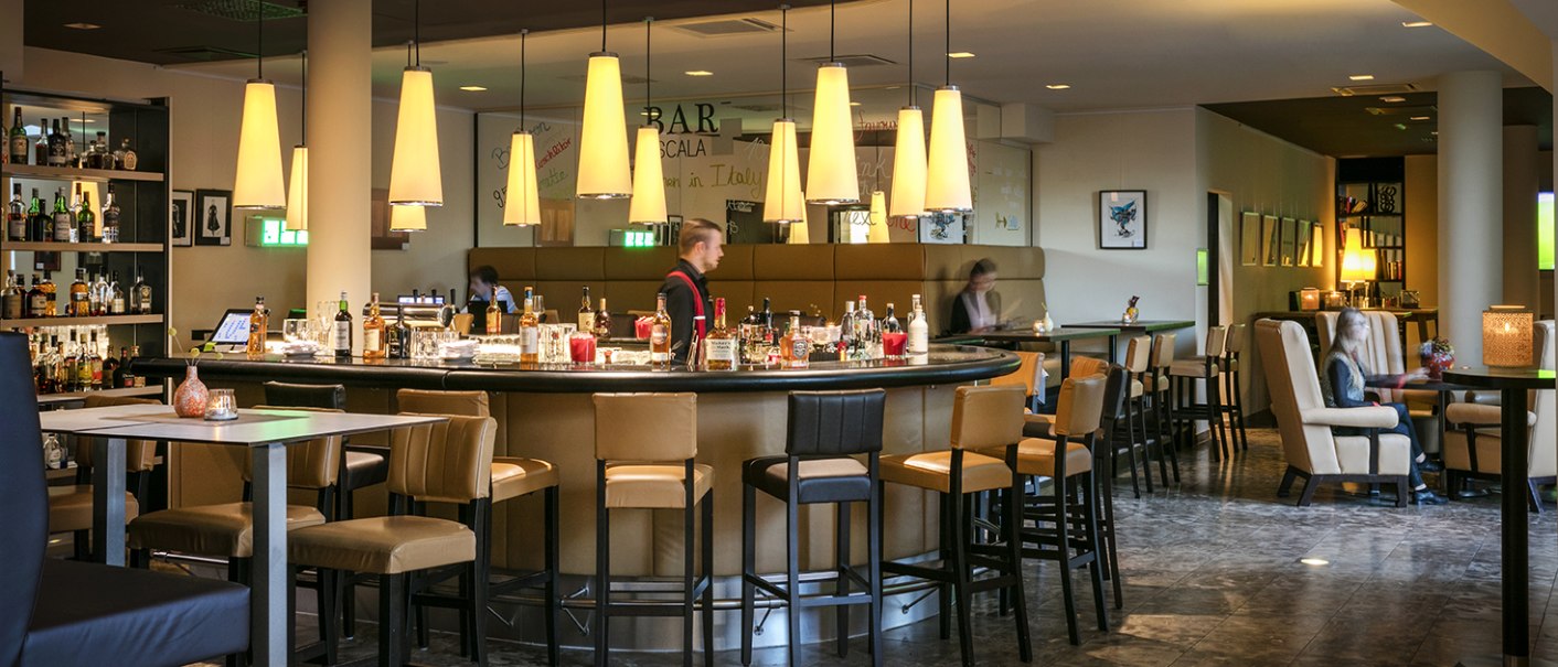 Elegante Hotelbar mit runder Theke, Barhockern und gedämpfter Beleuchtung. Gäste sitzen an Tischen, während ein Barkeeper hinter der Theke arbeitet., © Arcotel Hotels