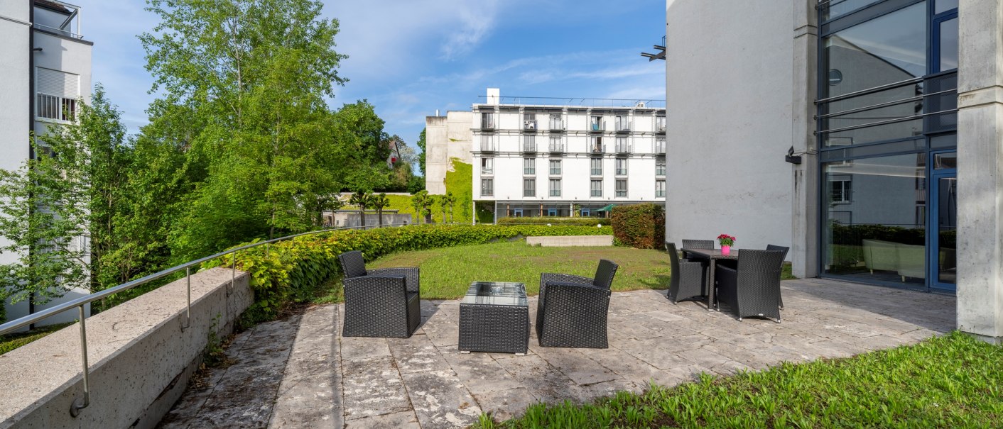 Moderne Terrasse mit Rattanmöbeln, Glasfenster und grüner Umgebung. Im Hintergrund sind Gebäude zu sehen., © Plaza Residence Stuttgart-Ditzingen Moderne Terrasse mit Rattanmöbeln, Glasfenster und grüner Umgebung. Im Hintergrund sind Gebäude zu sehen., © Plaza Residence Stuttgart-Ditzingen