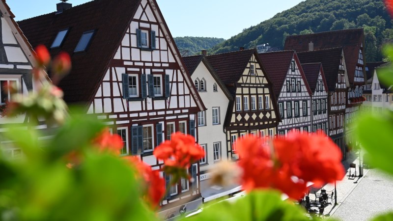 Fachwerkhäuser in einer Reihe, mit roten Blumen im Vordergrund. Im Hintergrund sind bewaldete Hügel zu sehen., © Bad Urach Tourismus Fachwerkhäuser in einer Reihe, mit roten Blumen im Vordergrund. Im Hintergrund sind bewaldete Hügel zu sehen., © Bad Urach Tourismus