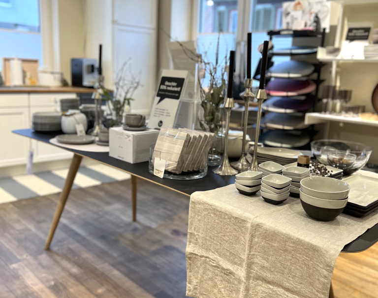 Ein Tisch in einem Laden mit Geschirr, Schalen, Kerzenhaltern und Vasen. Der Raum hat Holzboden und weiße Wände mit Regalen., © schöner laden