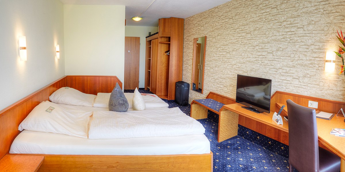 Ein gemütliches Doppelzimmer mit Holzmöbeln, Doppelbett, Schreibtisch, Fernseher und Koffer. Die Wände sind teils mit Steinoptik gestaltet., © Kemnater Hof GmbH