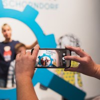 Eine Person fotografiert drei Kinder durch einen blauen, zahnradförmigen Rahmen mit einem Smartphone. Im Hintergrund sind Zeichnungen zu sehen., © Danijel Grbic