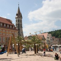Sandspielplatz mit Nestschaukel, Palmen und vielen Menschen. Im Hintergrund Häuser der Stadt und der Johannisturm., © Foto: Cornelia Steinbach