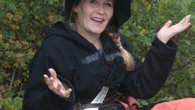 Eine Frau in mittelalterlicher Kleidung mit einem schwarzen Hut und einem Schwert posiert lächelnd im Wald., © Copyright: Stefanie Keller Eine Frau in mittelalterlicher Kleidung mit einem schwarzen Hut und einem Schwert posiert lächelnd im Wald., © Copyright: Stefanie Keller