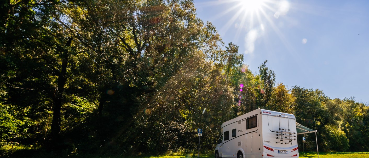Ein Wohnmobil steht auf einem Stellplatz im Gr&uuml;nen, umgeben von B&auml;umen. Die Sonne scheint hell am blauen Himmel., &copy; SMG, Thomas Niederm&uuml;ller