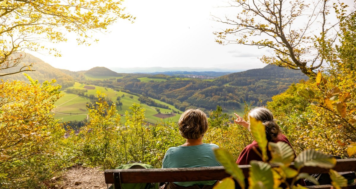 Zwei Personen sitzen auf einer Bank und genießen den Ausblick auf eine herbstliche Landschaft mit Hügeln und Feldern., © Foto: Mario Klaiber Zwei Personen sitzen auf einer Bank und genießen den Ausblick auf eine herbstliche Landschaft mit Hügeln und Feldern., © Foto: Mario Klaiber