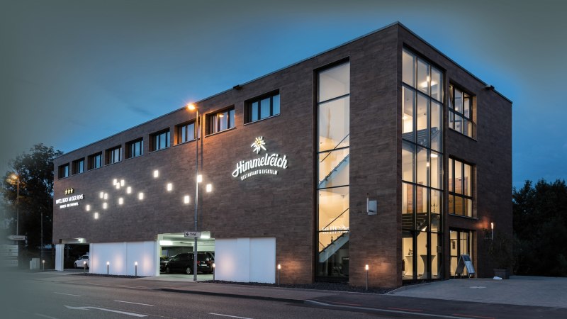 Modernes Hotelgebäude bei Nacht, beleuchtet mit großen Fenstern und Schildern. "Himmelreich Restaurant & Eventlocation" ist sichtbar., © Hotel Reich an der Rems
