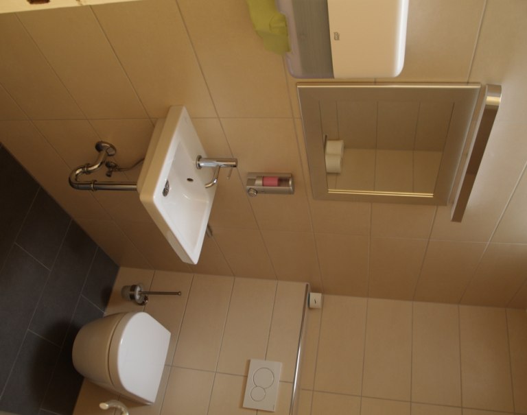 Ein kleines Badezimmer mit einem Waschbecken, einem Spiegel und einer Toilette. Die Wände sind beige gefliest., © TOMAS Ein kleines Badezimmer mit einem Waschbecken, einem Spiegel und einer Toilette. Die Wände sind beige gefliest., © TOMAS