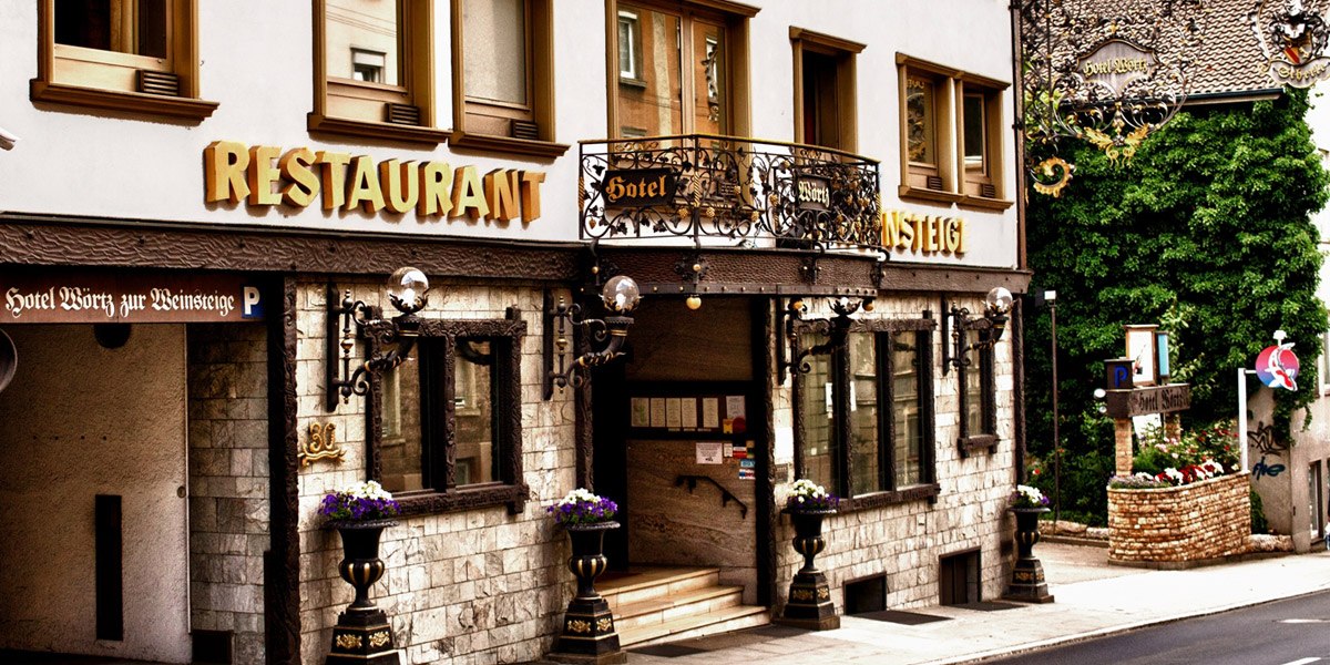 Eingang des Hotels Zur Weinsteige mit dekorativen Elementen, großen Fenstern und Restaurantbeschilderung. Blumen schmücken den Eingangsbereich., © Hotel Zur Weinsteige