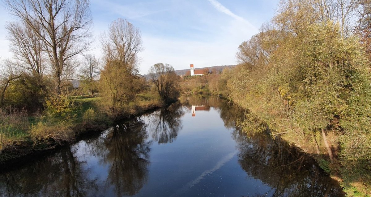 Ein ruhiger Fluss, gesäumt von kahlen Bäumen, spiegelt den blauen Himmel wider. Im Hintergrund ist eine Kirche mit rotem Dach zu sehen., © Kulturamt Plochingen Ein ruhiger Fluss, gesäumt von kahlen Bäumen, spiegelt den blauen Himmel wider. Im Hintergrund ist eine Kirche mit rotem Dach zu sehen., © Kulturamt Plochingen