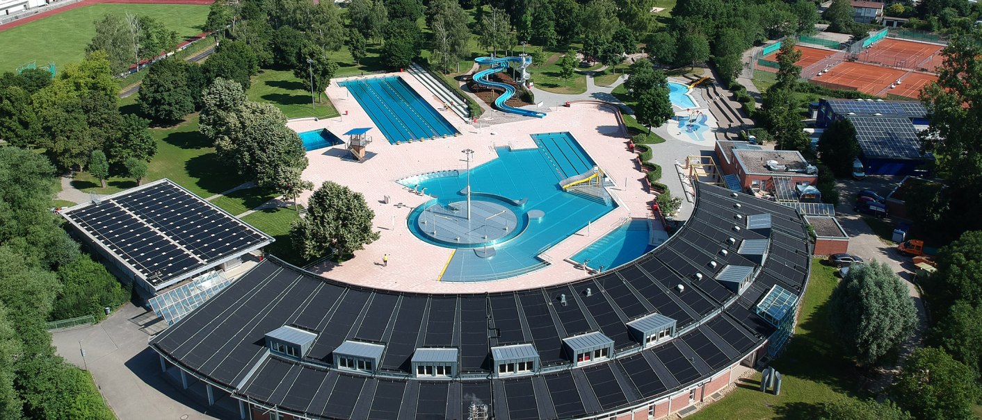 Luftaufnahme eines Freibads mit Becken, Rutschen und Solarzellen auf dem Dach. Umgeben von Bäumen und Sportplätzen., © Stadt Leonberg