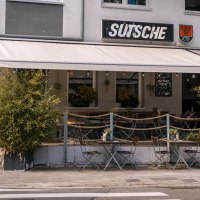 Café 'Sutsche' mit Außenbereich, Tischen und Stühlen auf dem Gehweg, umgeben von Pflanzen., © SMG, Sarah Schmid Café 'Sutsche' mit Außenbereich, Tischen und Stühlen auf dem Gehweg, umgeben von Pflanzen., © SMG, Sarah Schmid
