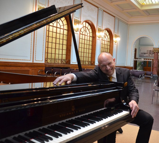 Ein Mann in einem Anzug lehnt sich lächelnd an einen Steinway-Flügel in einem eleganten Raum mit hohen Decken und verzierten Fenstern., © Tim Kandel Ein Mann in einem Anzug lehnt sich lächelnd an einen Steinway-Flügel in einem eleganten Raum mit hohen Decken und verzierten Fenstern., © Tim Kandel