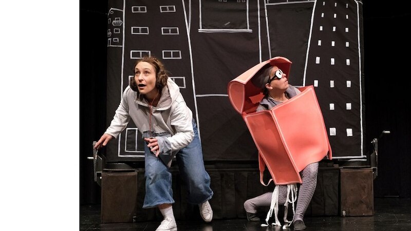 Zwei Personen auf einer B&uuml;hne, eine in Alltagskleidung, die andere in einem roten Kost&uuml;m, vor einem Hintergrund mit gezeichneten Geb&auml;uden., &copy; Kulturb&uuml;ro Schw&auml;bisch Gm&uuml;nd