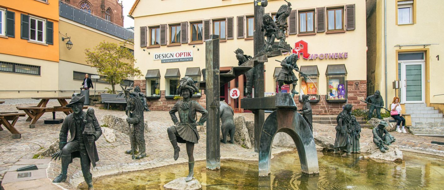 Der Narrenbrunnen in Weil der Stadt zeigt Bronzefiguren von Narren in verschiedenen Posen. Im Hintergrund sind Geschäfte und eine Apotheke zu sehen., © Stuttgart-Marketing GmbH, Sarah Schmid Der Narrenbrunnen in Weil der Stadt zeigt Bronzefiguren von Narren in verschiedenen Posen. Im Hintergrund sind Geschäfte und eine Apotheke zu sehen., © Stuttgart-Marketing GmbH, Sarah Schmid