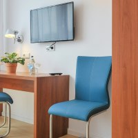 Ein schlichtes Hotelzimmer mit Schreibtisch, zwei blauen Stühlen, einem Fernseher an der Wand und einem Kleiderschrank., © Gästehaus ABZ Stuttgart