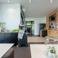 Moderne Hotel-Lounge mit Holzwänden, Snackbereich und Tisch mit Glasglocke. Zeitung und Kaffeemaschine sichtbar. Helle, einladende Atmosphäre., © a2 HOTELS