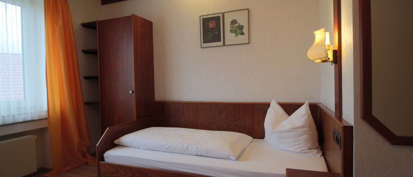 Ein gemütliches Einzelzimmer mit Holzmöbeln, einem Einzelbett, orange Vorhängen und zwei Wandbildern. Eine Lampe beleuchtet den Raum., © TOMAS Ein gemütliches Einzelzimmer mit Holzmöbeln, einem Einzelbett, orange Vorhängen und zwei Wandbildern. Eine Lampe beleuchtet den Raum., © TOMAS