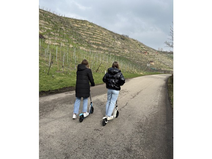 Zwei Personen mit Rollern fahren auf einer Stra&szlig;e entlang eines Weinbergs. Sie tragen warme Jacken und Jeans. Der Himmel ist bew&ouml;lkt., &copy; Cool-Tours StattReisen