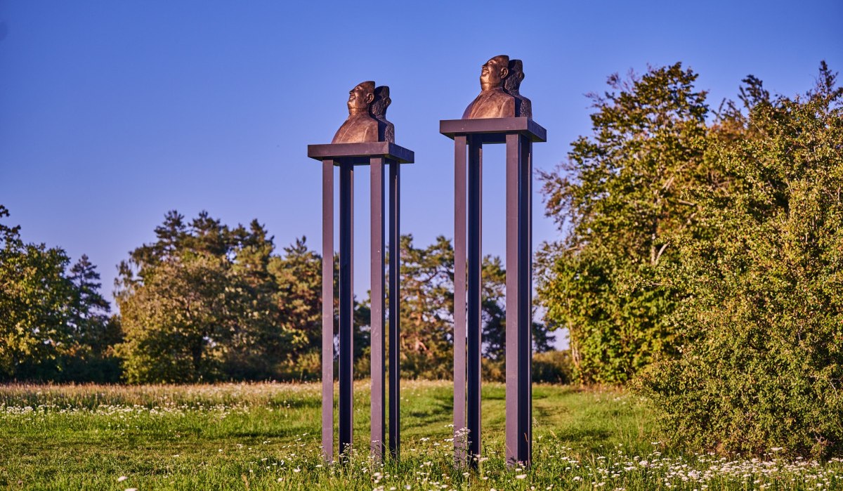 Zwei Büsten auf hohen, schlanken Säulen stehen auf einer grünen Wiese, umgeben von Bäumen und blauem Himmel., © Natur.Nah. Schönbuch & Heckengäu Zwei Büsten auf hohen, schlanken Säulen stehen auf einer grünen Wiese, umgeben von Bäumen und blauem Himmel., © Natur.Nah. Schönbuch & Heckengäu