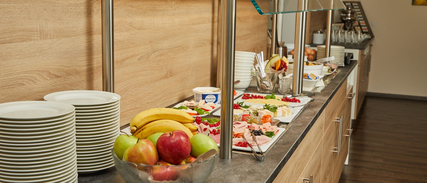 Ein Frühstücksbuffet mit Obstschale, Aufschnittplatten und gestapelten Tellern auf einer Theke., © TOMAS