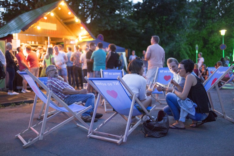 Menschen sitzen in Liegestühlen bei einem Fest. Im Hintergrund ein beleuchteter Stand, viele Besucher stehen an. Entspannte Abendstimmung., © D. Jüptner Menschen sitzen in Liegestühlen bei einem Fest. Im Hintergrund ein beleuchteter Stand, viele Besucher stehen an. Entspannte Abendstimmung., © D. Jüptner