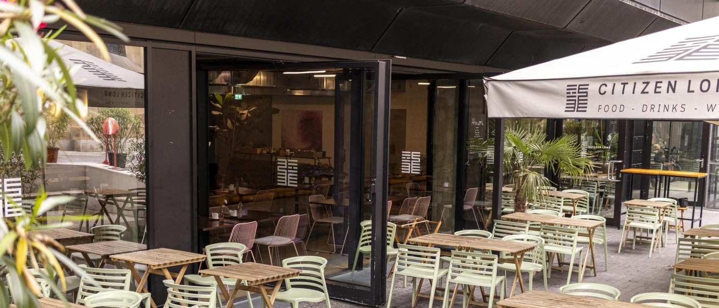 Außenbereich eines Cafés mit Holztischen und grünen Stühlen. Eine Markise trägt den Schriftzug 'Citizen Long'. Pflanzen dekorieren den Bereich., © SMG, Sarah Schmid