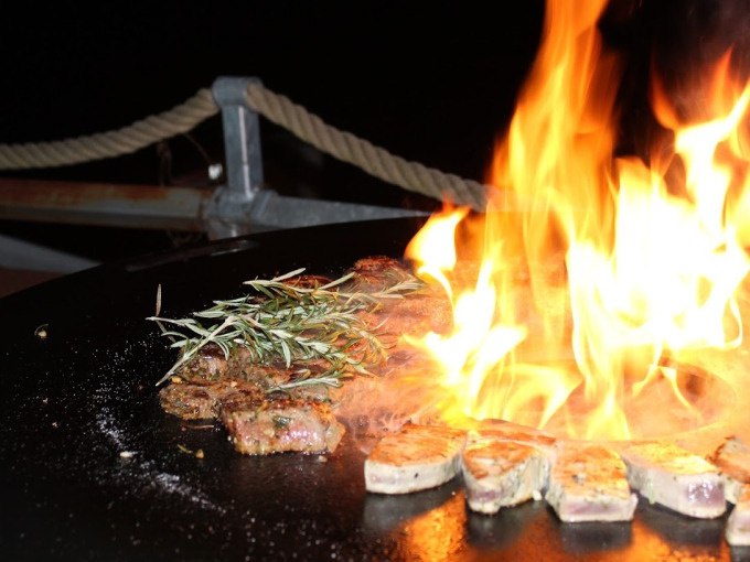 Ein Grill mit brennendem Feuer, Fleischstücken und Rosmarinzweigen im Dunkeln. Im Hintergrund ein Seilgeländer., © Cool-Tours StattReisen