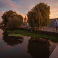 Ein Fluss bei Sonnenuntergang mit B&auml;umen und einem Turm im Hintergrund. Menschen spazieren entlang des Ufers, w&auml;hrend der Himmel in warmen Farben leuchtet., &copy; WTM GmbH Waiblingen