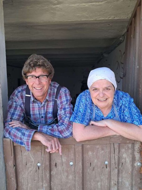 Zwei Personen lehnen sich lächelnd über eine Holztür. Eine Person trägt ein kariertes Hemd, die andere ein blaues Kopftuch und Hemd., © SBEntertainment GmbH & Co. KG Zwei Personen lehnen sich lächelnd über eine Holztür. Eine Person trägt ein kariertes Hemd, die andere ein blaues Kopftuch und Hemd., © SBEntertainment GmbH & Co. KG