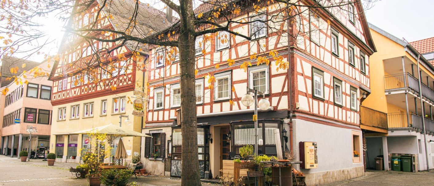 Fachwerkhäuser in der Altstadt von Künzelsau, umgeben von herbstlichen Bäumen. Die Sonne scheint durch die Äste und beleuchtet die Szene., © Stuttgart-Marketing GmbH, Sarah Schmid