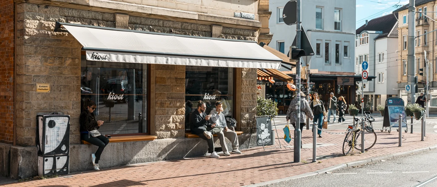 Straßenszene vor dem Café Kiost mit Passanten, Fahrrädern und Sonnenschutz an einer belebten Straßenecke., © SMG, Sarah Schmid Straßenszene vor dem Café Kiost mit Passanten, Fahrrädern und Sonnenschutz an einer belebten Straßenecke., © SMG, Sarah Schmid