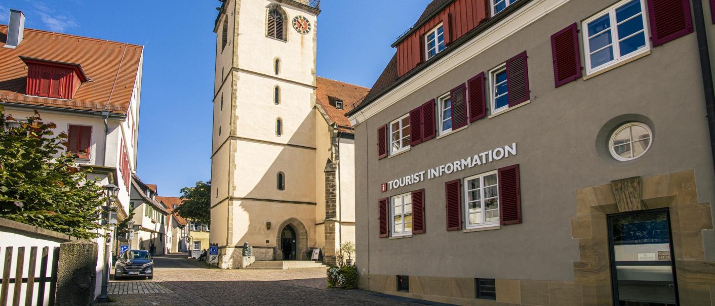 Gebäude mit der Aufschrift 'Tourist Information' neben einer Kirche mit Turm und roten Fensterläden, bei sonnigem Wetter., © Stuttgart-Marketing GmbH, Sarah Schmid