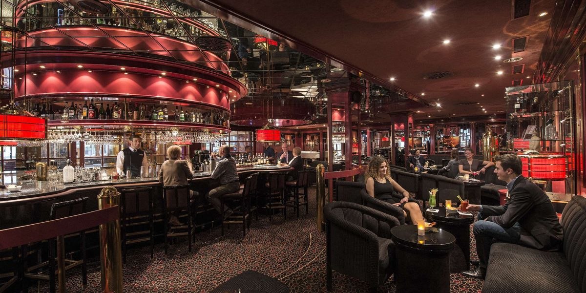 Elegante Bar mit rotem Dekor, Menschen sitzen an Tischen und der Theke, gedämpfte Beleuchtung., © Maritim Hotelgesellschaft mbH