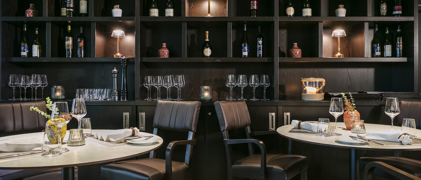 Elegantes Restaurant mit gedeckten Tischen, Weingl&auml;sern und einem Regal voller Weinflaschen und Dekorationen im Hintergrund., &copy; Arcotel Hotels
