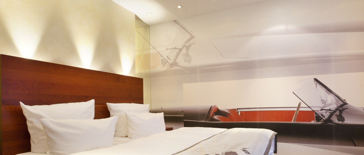 Modernes Hotelzimmer mit großem Bett, Holzkopfteil und Wandbild eines Autos. Dezente Beleuchtung und weiße Bettwäsche., © Ringhotel Adler Asperg