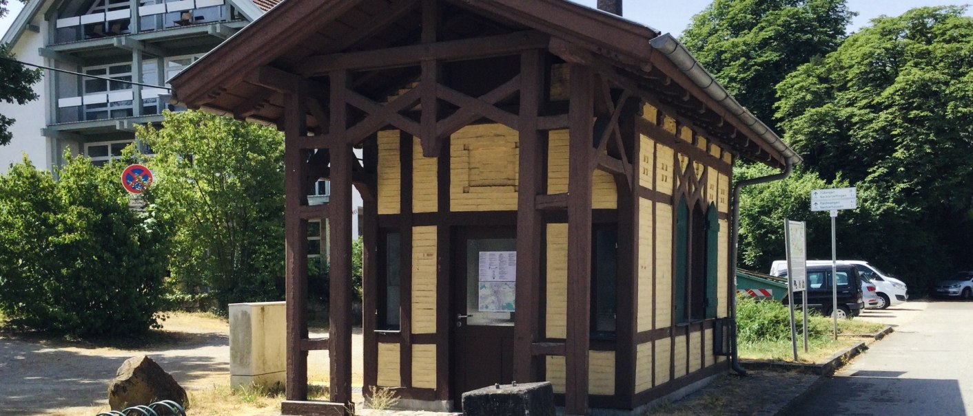 Kleines Fachwerkgebäude am Neckartalradweg, umgeben von Bäumen und einem modernen Gebäude. Ein Infopunkt mit Schildern und Fahrradständer., © J. Tubolzer
