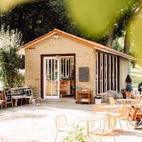 Kleines Café mit Steinwänden und großen Fenstern, umgeben von Bäumen. Orangefarbene Stühle und Tische auf der Terrasse, einladende Atmosphäre., © L. Prestle/ Documenting Love Kleines Café mit Steinwänden und großen Fenstern, umgeben von Bäumen. Orangefarbene Stühle und Tische auf der Terrasse, einladende Atmosphäre., © L. Prestle/ Documenting Love
