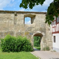 Historische Stadtmauer mit kleinem Torbogen und leicht nach links versetztem "Fenster", rechts an die Mauer anschlie&szlig;end ein Fachwerkhaus mit roten Holzbalken. Im Vordergrund eine Wiese mit Str&auml;uchern., &copy; Petra Natzkowski