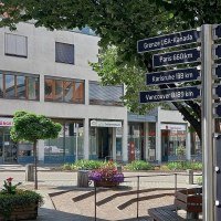 Wegweiser auf einem Platz mit Entfernungen zu Städten und Orten auf dem 49. Breitengrad, wie Paris und Vancouver. Im Hintergrund ein modernes, weißes Gebäude, © Petra Natzkowski Wegweiser auf einem Platz mit Entfernungen zu Städten und Orten auf dem 49. Breitengrad, wie Paris und Vancouver. Im Hintergrund ein modernes, weißes Gebäude, © Petra Natzkowski