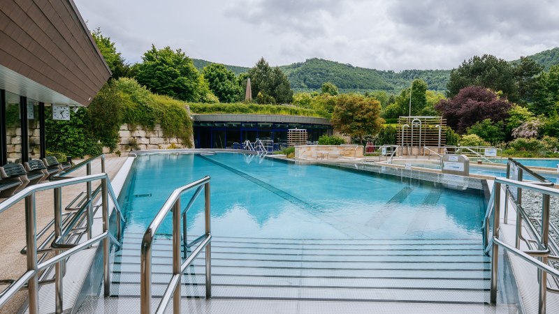 Ein Außenpool in einer Therme, umgeben von üppiger Vegetation und Hügeln im Hintergrund. Metallgeländer führen ins Wasser., © Thomas Niedermüller Ein Außenpool in einer Therme, umgeben von üppiger Vegetation und Hügeln im Hintergrund. Metallgeländer führen ins Wasser., © Thomas Niedermüller