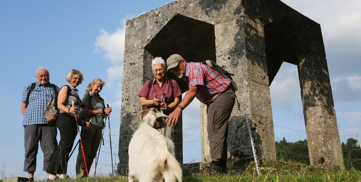 Wanderer stehen mit einem Hund vor einem steinernen Ehrenmal auf einer Wiese. Der Himmel ist blau mit einigen Wolken., © Bad Urach Tourismus
