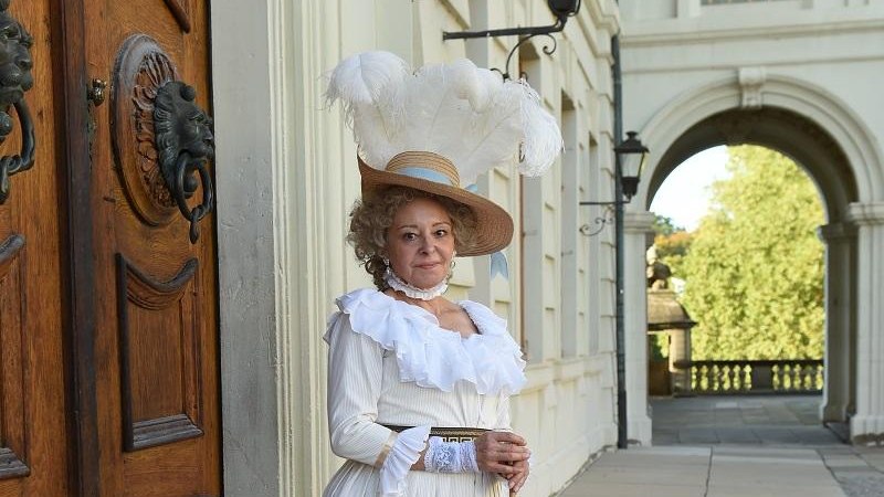 Eine Frau in einem eleganten, historischen Kost&uuml;m mit Hut und Feder steht vor einem Schloss. Sie h&auml;lt einen Sonnenschirm und l&auml;chelt in die Kamera., &copy; Fotograf: Reiner Pfisterer