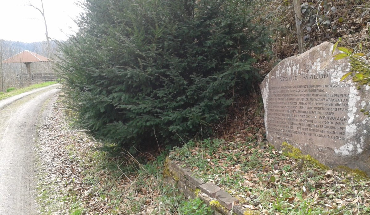 Ein Gedenkstein mit Inschrift steht am Rand eines Waldwegs. Ein großer Busch wächst daneben. Im Hintergrund ist ein Pavillon zu sehen., © Natur.Nah. Schönbuch & Heckengäu Ein Gedenkstein mit Inschrift steht am Rand eines Waldwegs. Ein großer Busch wächst daneben. Im Hintergrund ist ein Pavillon zu sehen., © Natur.Nah. Schönbuch & Heckengäu