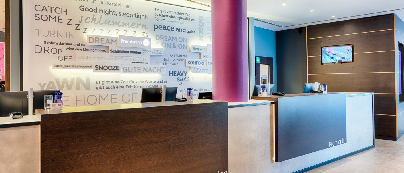 Moderne Hotelrezeption mit zwei Schreibtischen, lila Säule und Wand voller Schlafzitate. Premier Inn-Logo sichtbar., © Premier Inn Germany