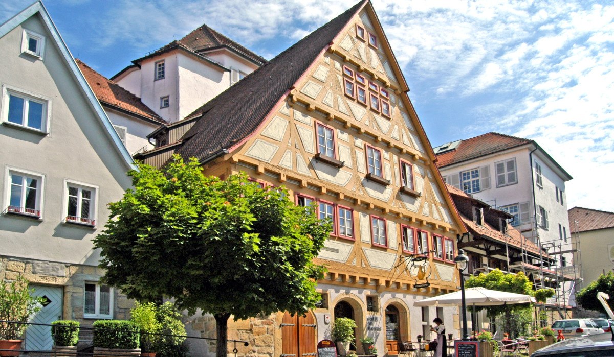 Fachwerkhaus in einer Altstadt mit Café und Sonnenschirmen. Menschen sitzen draußen, ein Baum spendet Schatten. Der Himmel ist blau mit wenigen Wolken., © Graphic Design Albig, GDA Waldenbuch